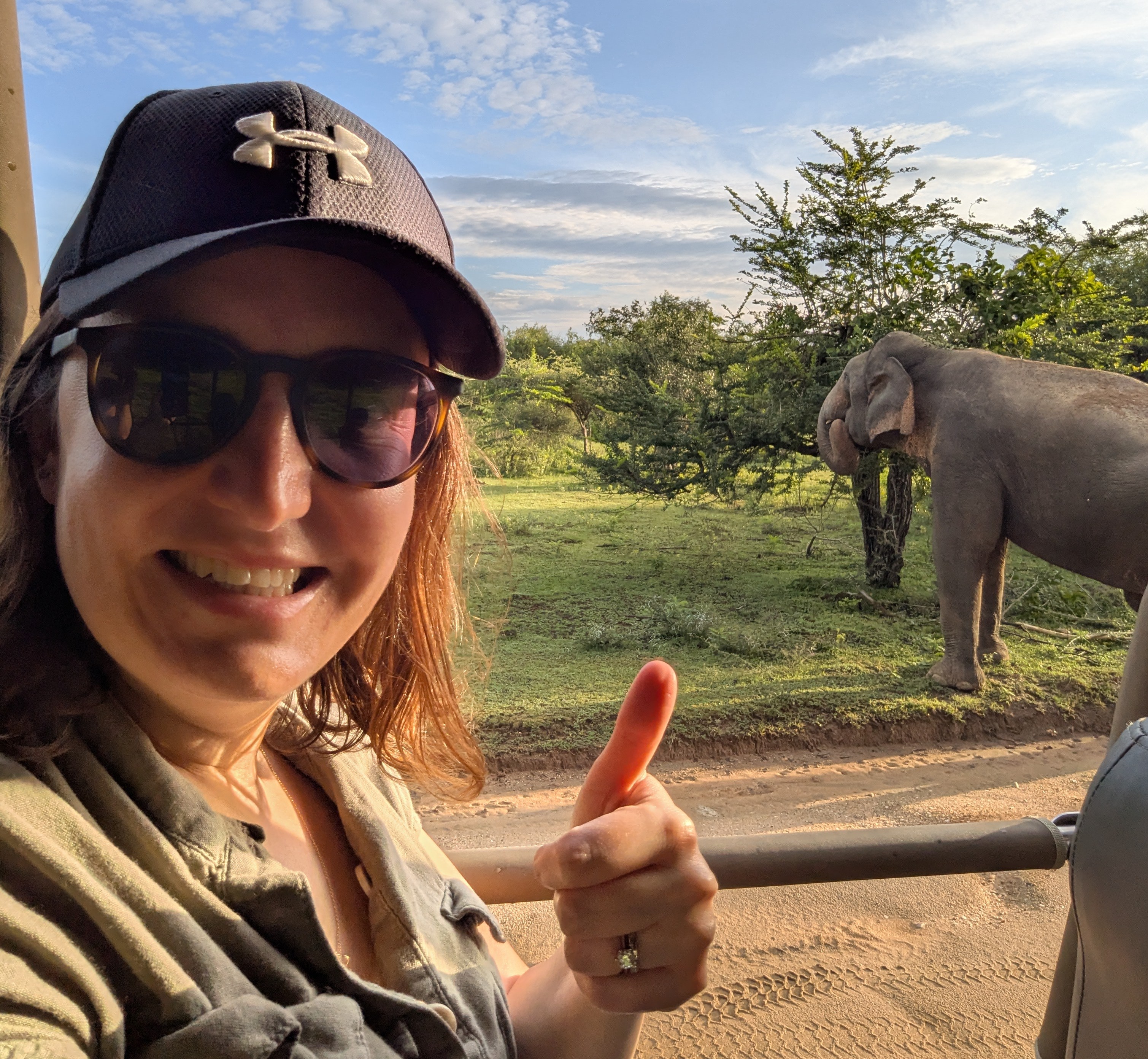 Woman giving thumbs up while on safari, with elephant in background.