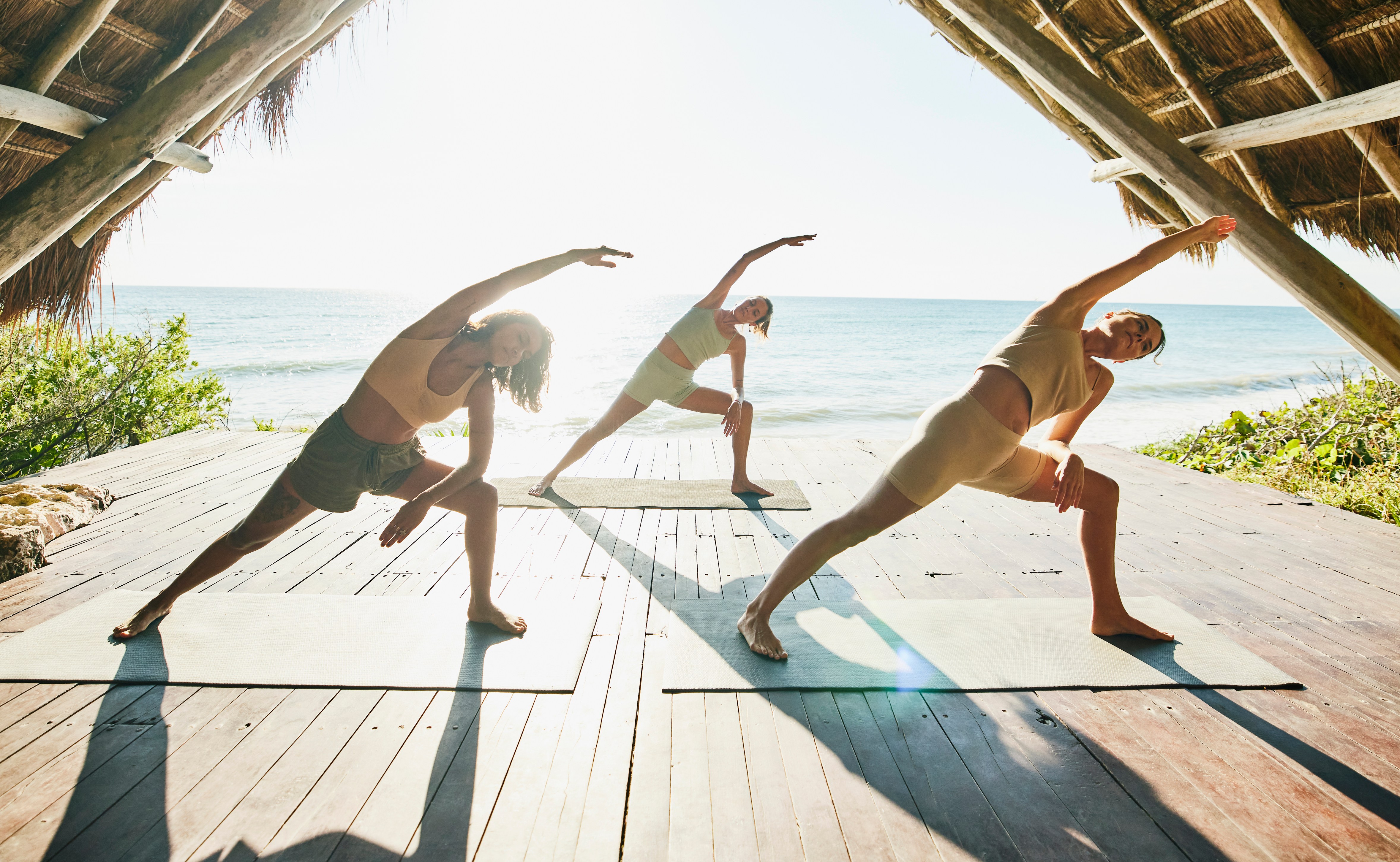 Three women practicing yoga in extended side angle pose on a beachside deck.