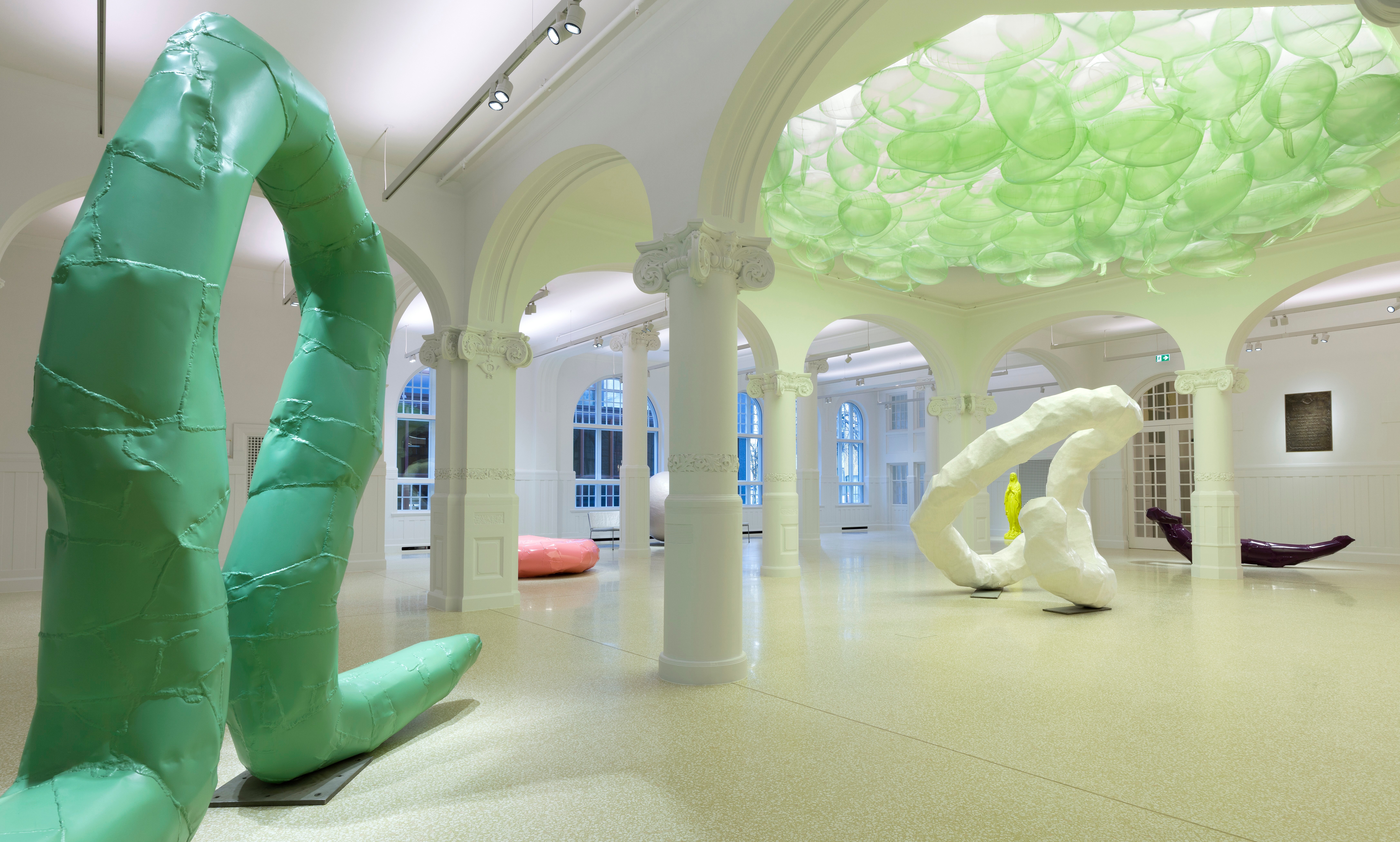 Interior view of an art gallery with several sculptures and a light green balloon installation on the ceiling.